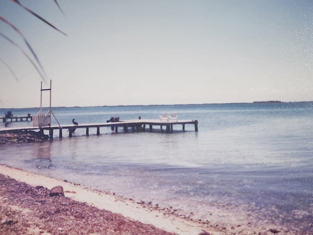 A calm beach scene featuring a wooden dock extending into the water with chairs and distant islands.
