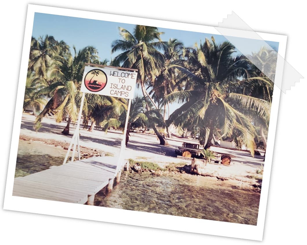 A sign reading "Welcome to Island Camps" stands on a dock surrounded by palm trees and sandy beach.