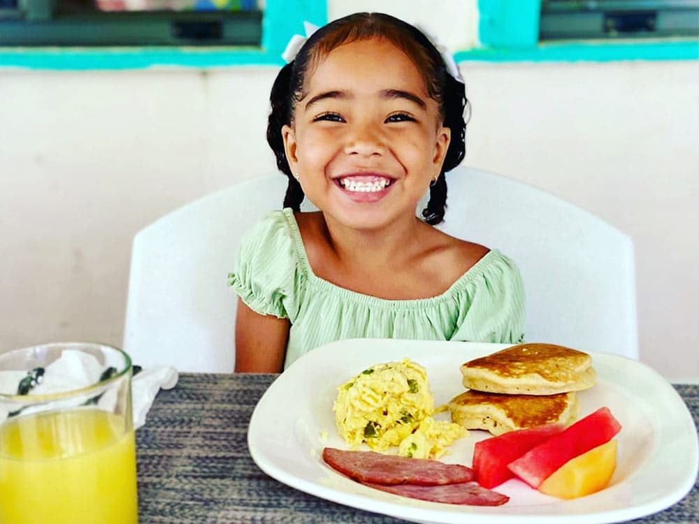 A smiling girl in a green dress sits at a table with a plate of breakfast, including scrambled eggs, pancakes, fruit, and a glass of juice.