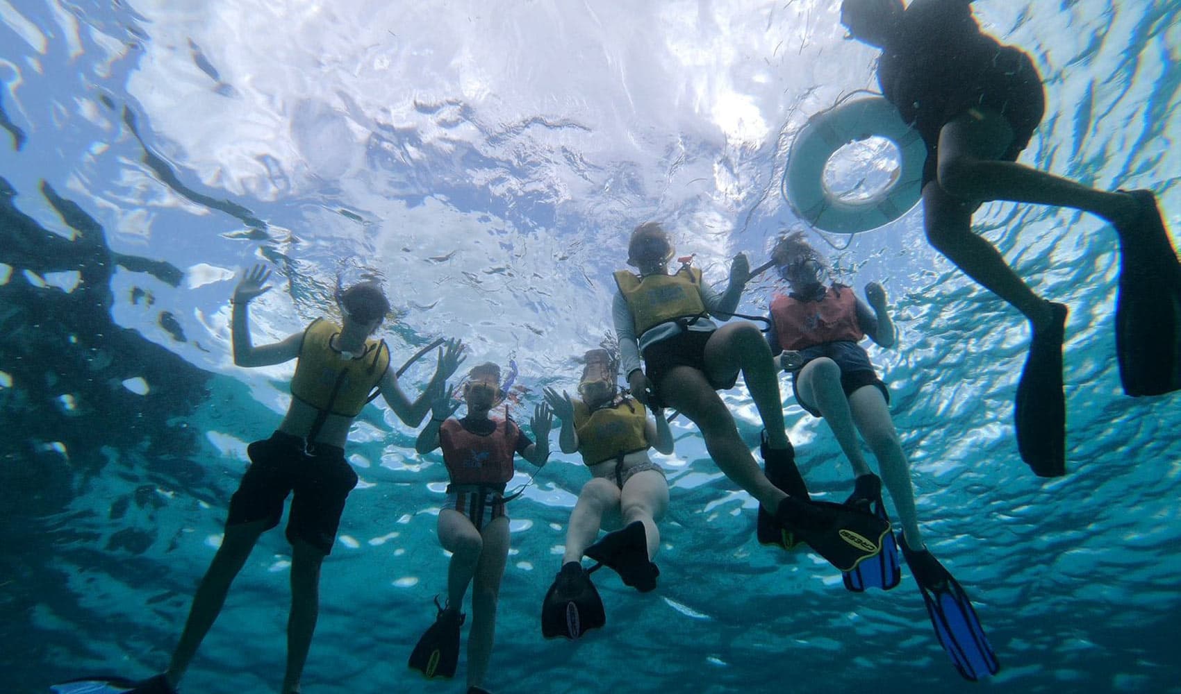 Six people in snorkel gear are submerged underwater, waving and enjoying the aquatic environment.