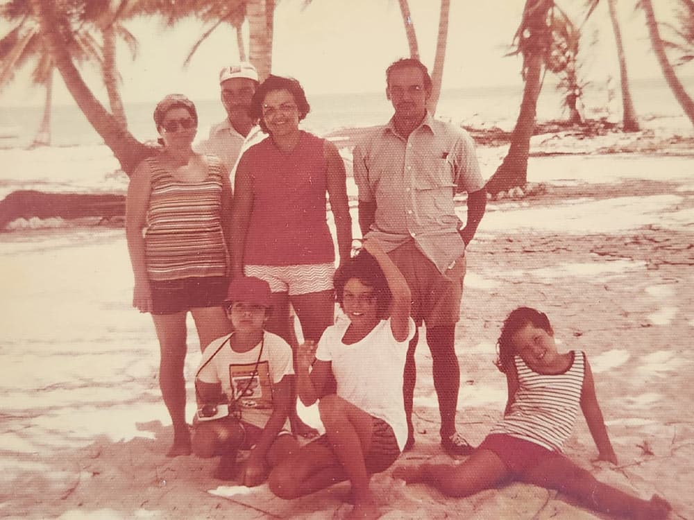 A group of seven people, including adults and children, poses together on a beach surrounded by palm trees.