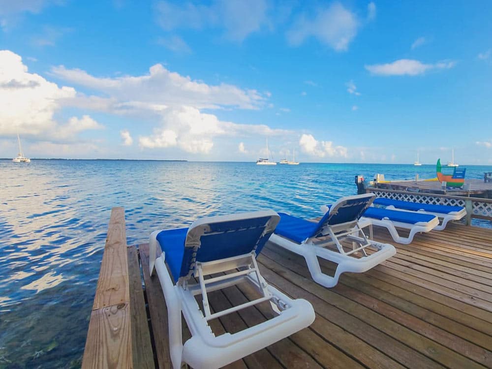 Lounge chairs on a wooden deck overlooking a calm sea and sailboats under a blue sky.