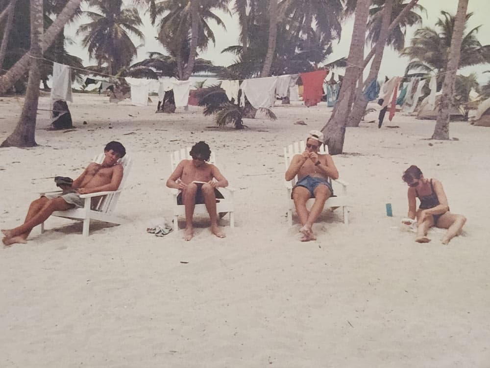 Four people relax on beach chairs on a sandy beach, with palm trees and laundry hanging in the background.